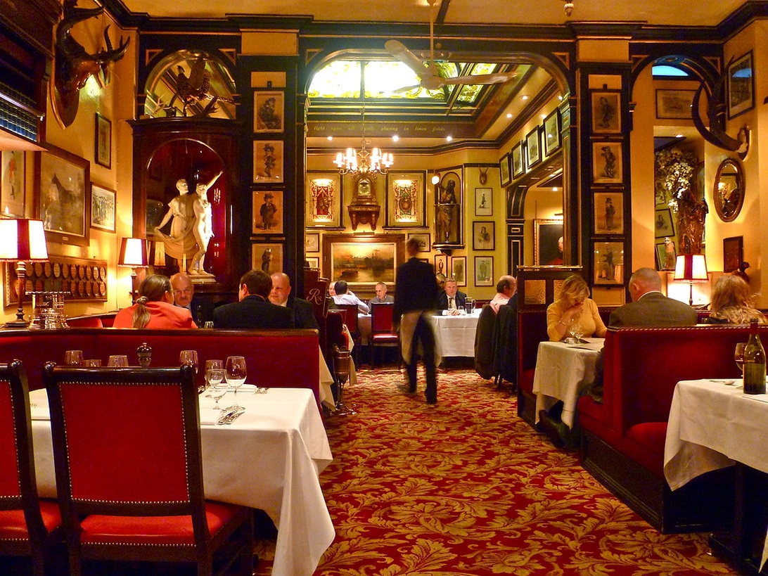 En klassisk, exklusiv restauranginteriör med rik dekor, röda stoppade säten, vita bordsdukar och utsmyckade väggdekorationer. Utrymmet har varm belysning och traditionella konstverk på väggarna.